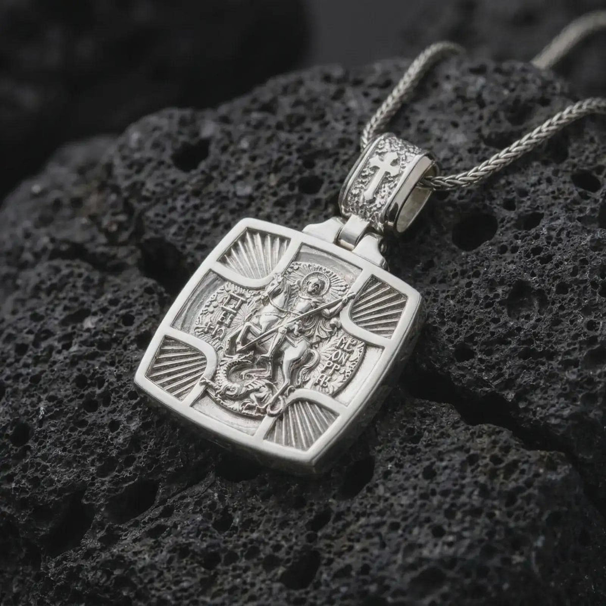 Polished Silver Saint George pendant displayed on black volcanic rock, highlighting the sculpted dragon, Greek inscriptions, and cross-shaped frame.