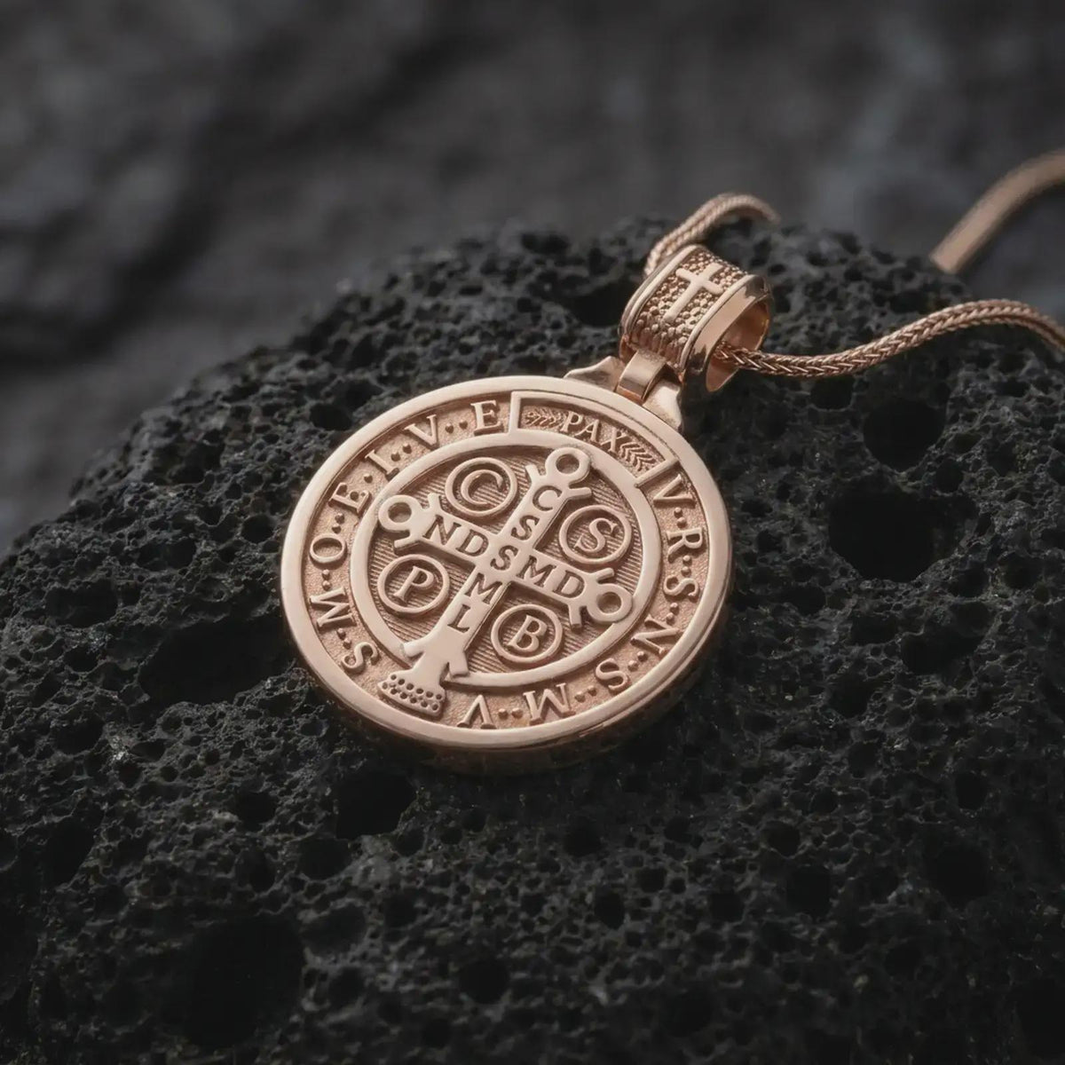Rose Gold Plated Saint Benedict religious pendant on black volcanic rock, showing the circular Benedict medal design with symbolic inscriptions.