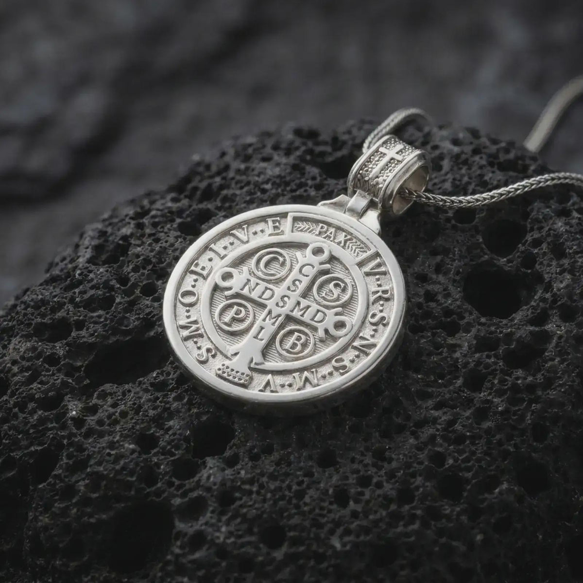 Polished Silver Saint Benedict medal necklace on black volcanic rock, featuring a round religious pendant with cross symbols and detailed lettering.