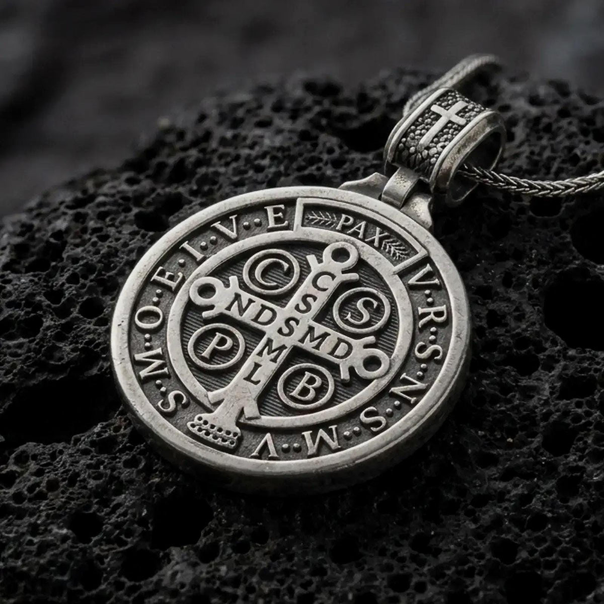 Oxidized Silver double sided Saint Benedict pendant on black volcanic rock, showing the Benedict medal cross with Latin initials and PAX inscription.
