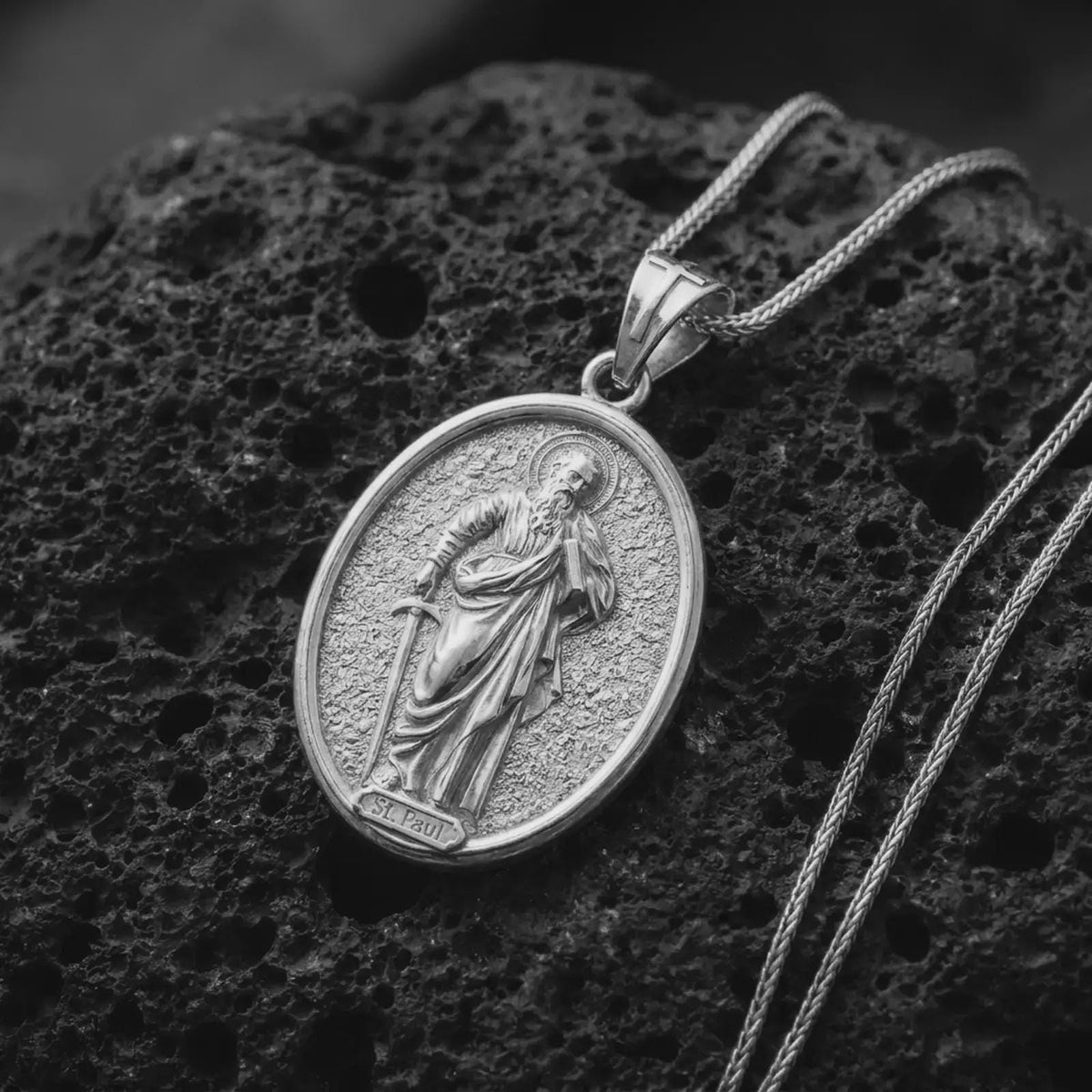 Polished Silver St Paul necklace on black volcanic rock, highlighting the oval religious medal, textured background, and Saint Paul inscription.