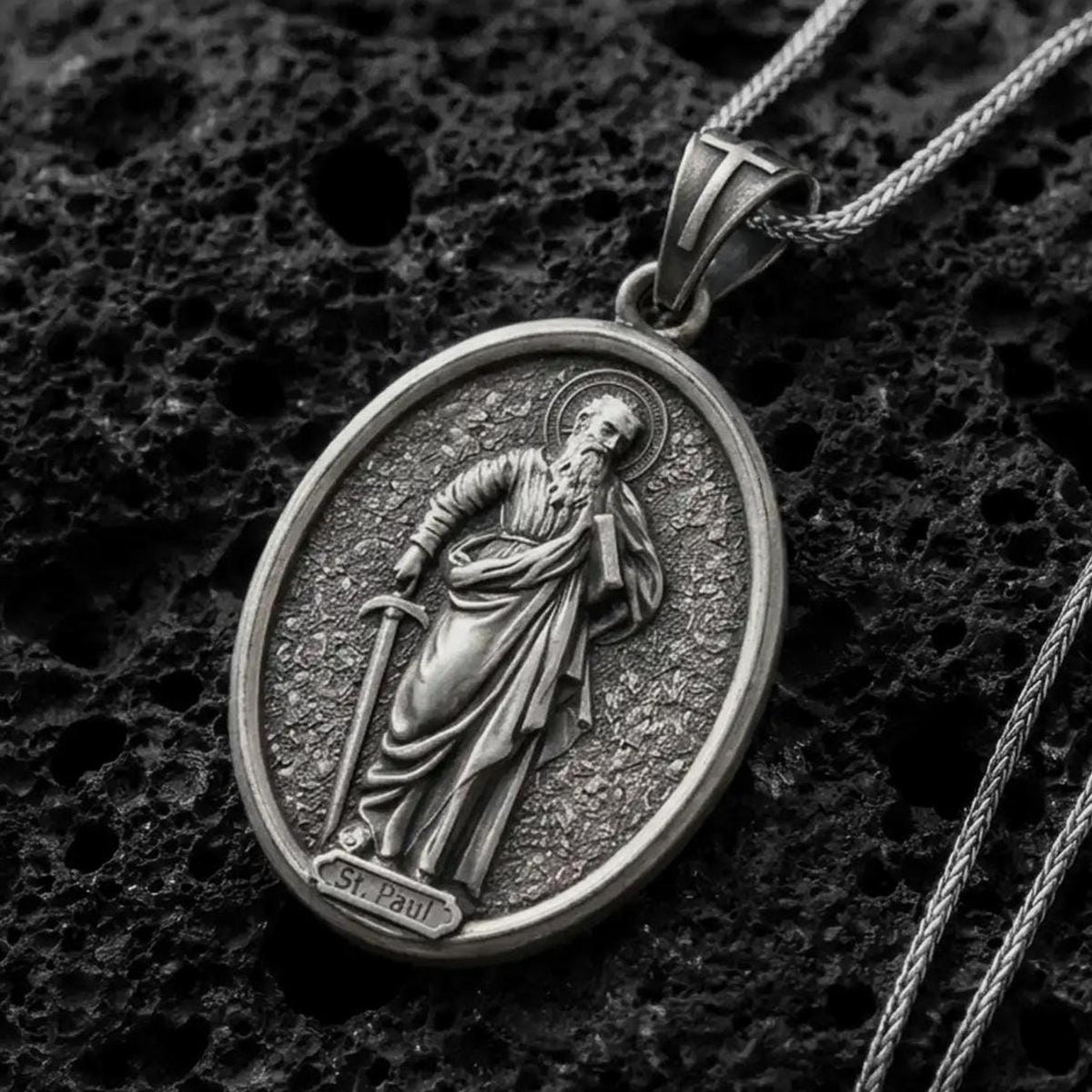 Oxidized Silver Saint Paul Apostle pendant on black volcanic rock, featuring the haloed saint holding a sword and book in detailed relief.