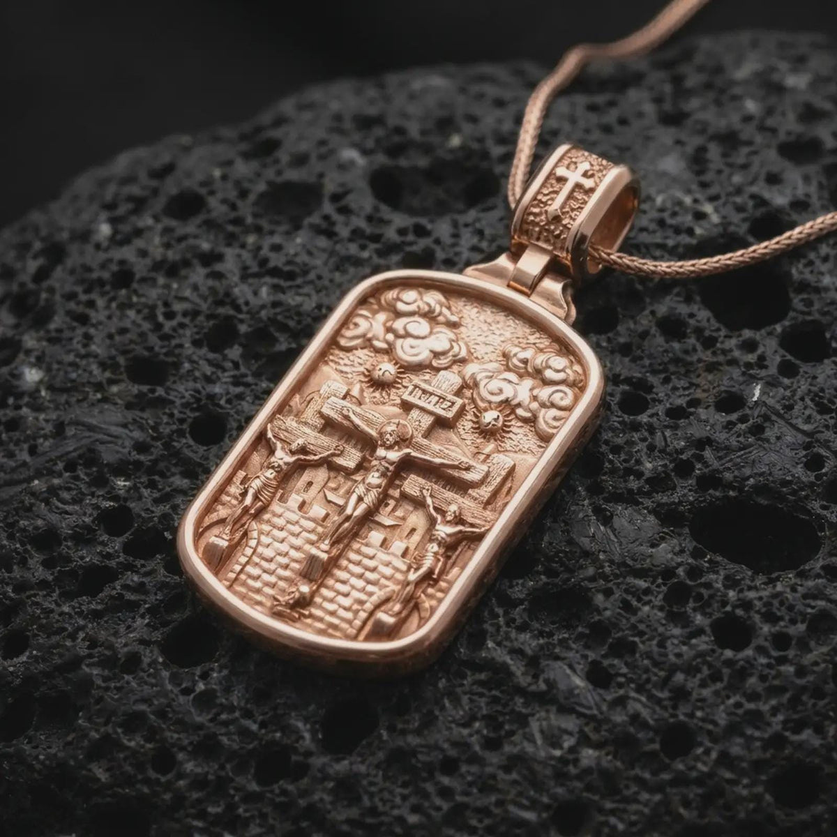 Rose Gold Plated Three Crosses Christian medal resting on black volcanic rock, showing the detailed crucifixion scene and cross-decorated bail.