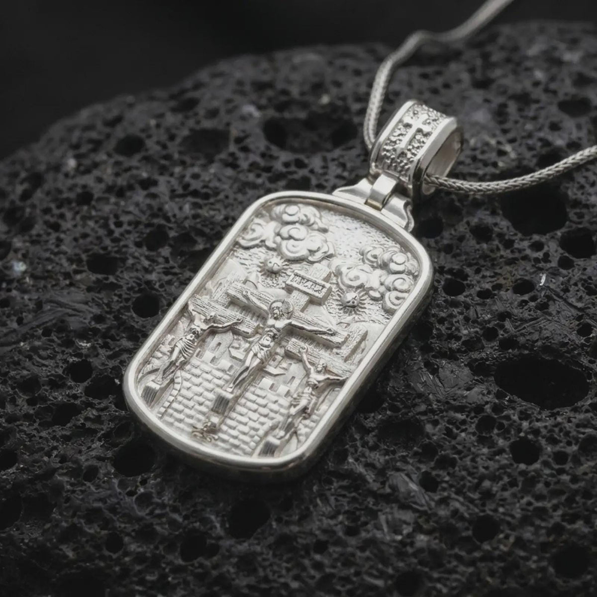 Polished Silver Calvary Crucifixion necklace on black volcanic rock, highlighting three sculpted crosses and detailed biblical figures.