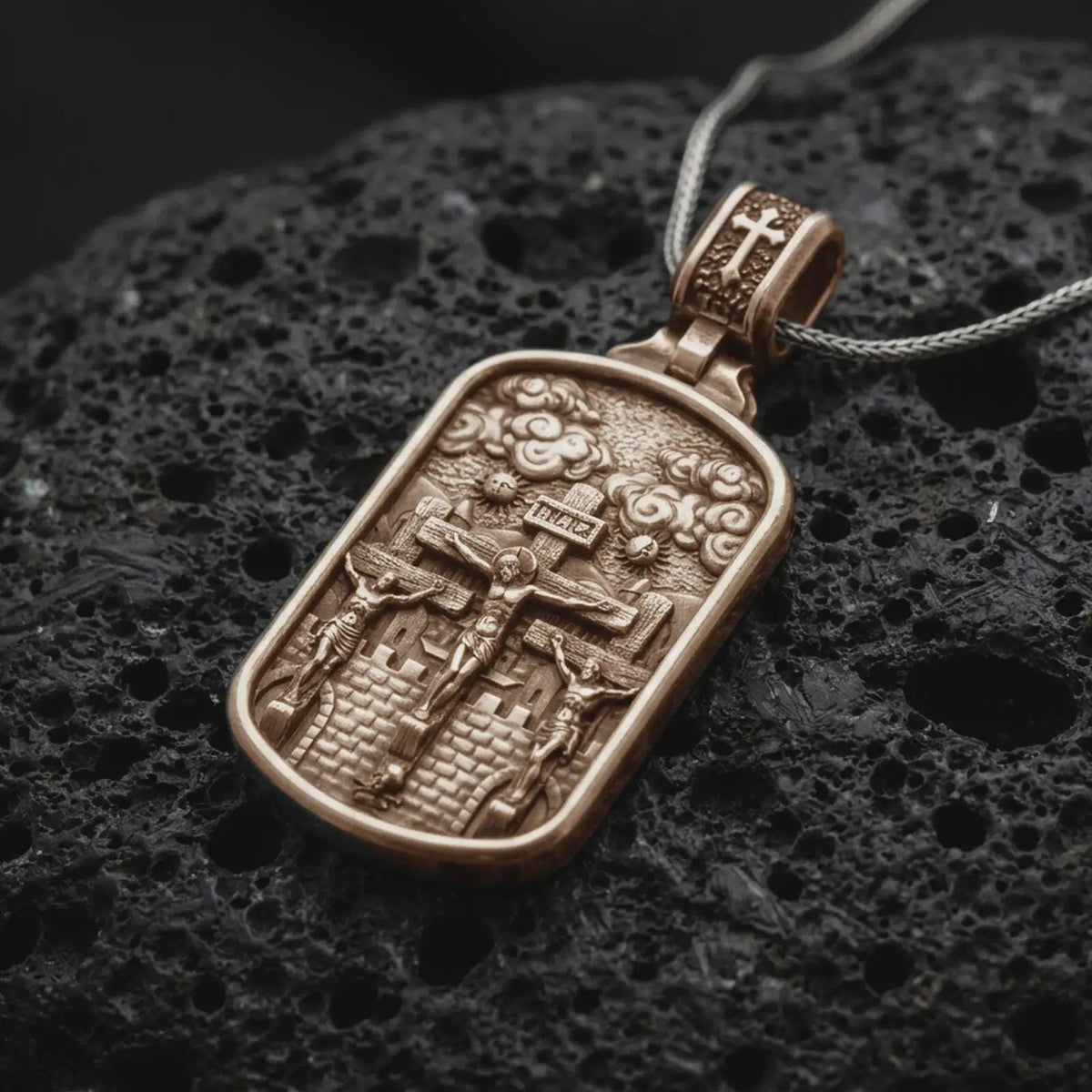 Bronze Three Crosses Crucifixion pendant on black volcanic rock, featuring Jesus Christ between two thieves at Calvary.