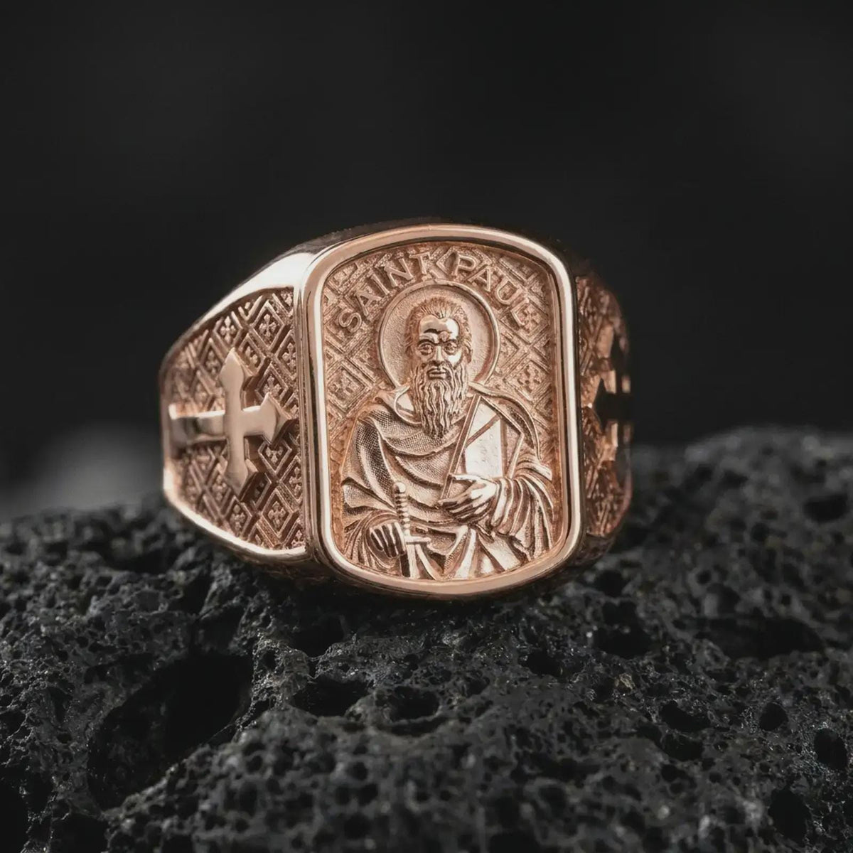 Rose Gold Plated Saint Paul Catholic signet ring resting on black volcanic rock, featuring sculpted robes, a traditional halo, and ornate cross engravings.