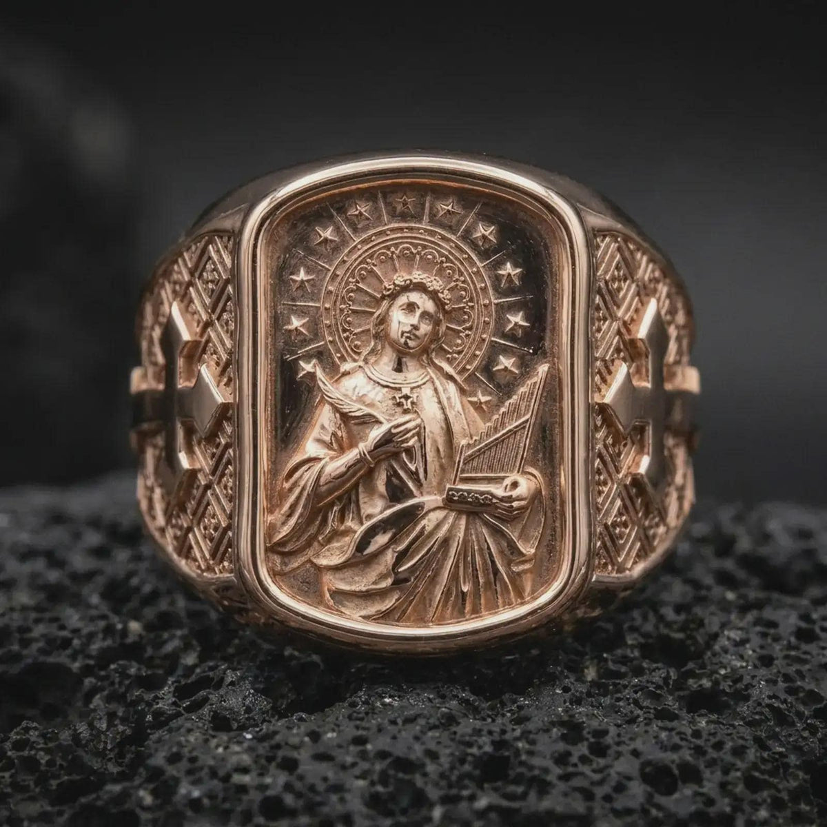 Rose Gold Plated Saint Cecilia religious signet ring resting on black volcanic rock, featuring flowing robes, musical symbolism, and decorative side crosses.