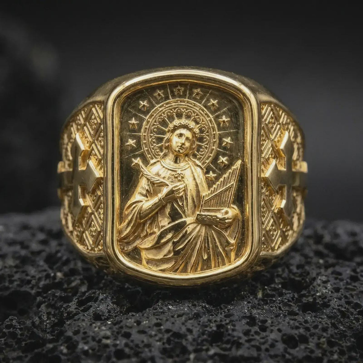 Gold Plated Saint Cecilia Catholic ring displayed on black volcanic rock, showcasing the ornate geometric band and raised cross motifs.